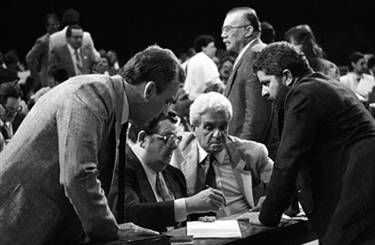 Foto preto e branco de homens conversando em torno de um papel