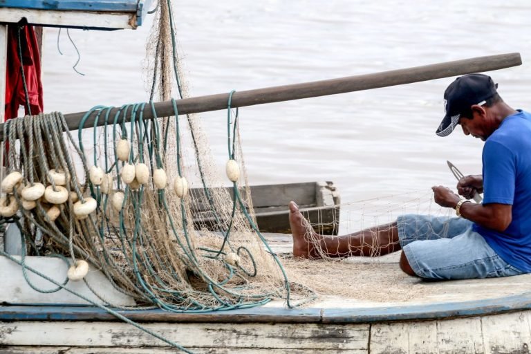 Pescador sentado num barco conserta a rede de pesca. Ele veste uma camisa azul e um boné preto