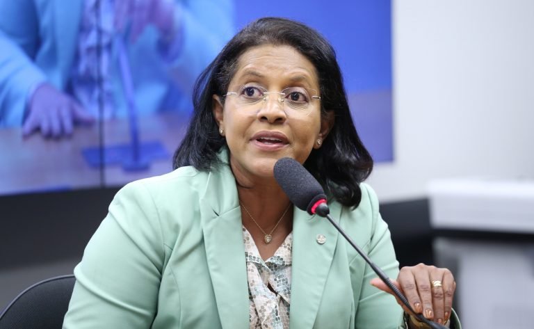 Deputada Gisela Simona fala ao microfone. Ela é uma mulher morena, com cabelo escuro e liso, usa óculo e veste um blazer verde claro