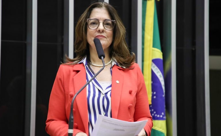 Deputada Rogéria Santos fala ao microfone. Ela é branca, usa óculos e um blazer laranja