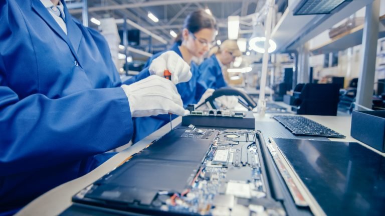 Close-up de um trabalhador fêmea da fábrica da eletrônica no casaco de trabalho azul que monta o cartão-matriz do portátil com uma chave de fenda. Instalação de fábrica de alta tecnologia com múltiplos funcionários.