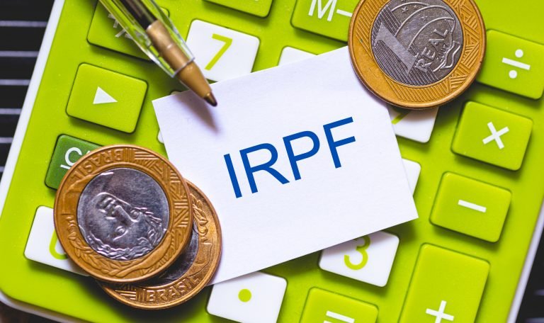 Imagem mostra moedas, uma caneta e um papel com a sigla IRPF (Imposto sobre a Renda da Pessoa Física) em cima de uma calculadora