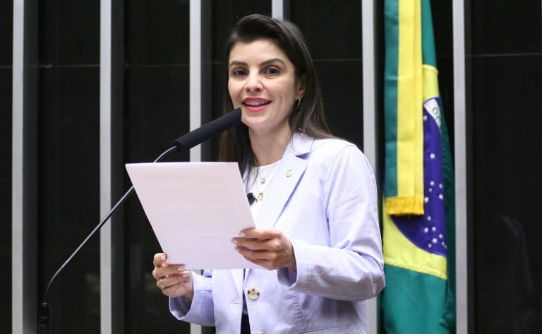 Deputada Marussa Boldrin fala ao microfone. Ela é branca, tem cabelo comprido, liso e escuro; veste um blazer claro e segura um papel. Ao fundo, à direita, há uma bandeira do Brasil num mastro
