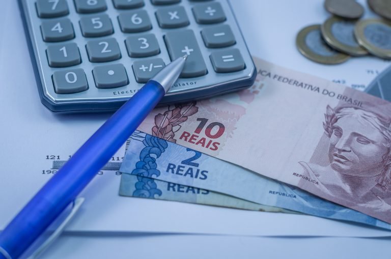 Imagem mostra calculadora, caneta, cédulas e moedas em cima de papéis