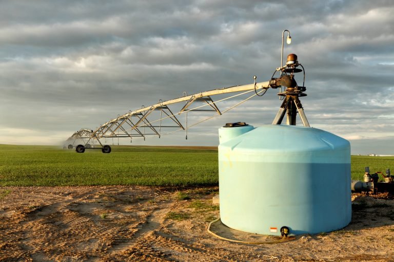 Campo agrícola com sistema de irrigação. Em primeiro plano, há um tanque de fertigação ligado à estrutura usada para distribuir água e nutrientes pela plantação.